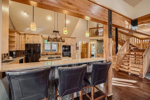 A modern kitchen with wooden cabinets and a large island. Three black barstools line the island, and a wooden staircase is visible in the background. Hanging lights illuminate the kitchen.