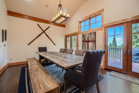 A rustic dining room with a large wooden table, leather chairs, a bench, and ski decor on the wall. The room has high ceilings, large windows, and glass doors leading to a wooden deck with forest views.